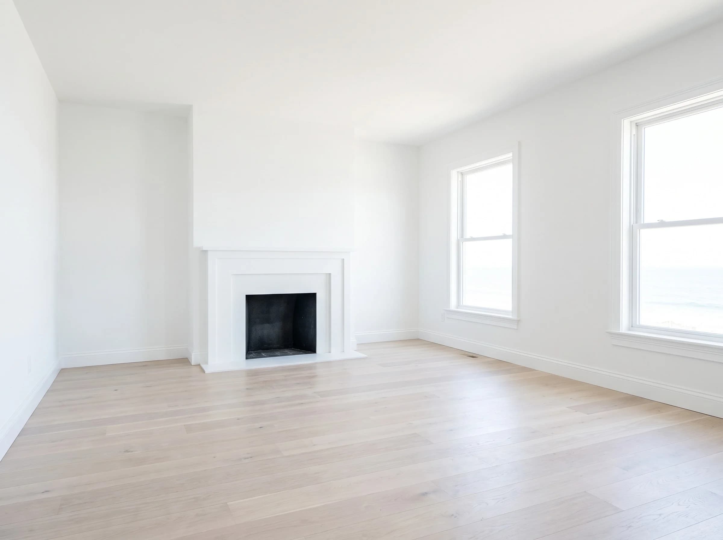 Empty living room with a white fireplace, two tall windows and pale wood floors.